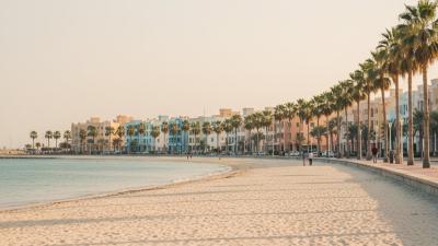 Ajman Corniche at golden hour: a soft white-sand beach meets the calm turquoise Arabian Gulf, with a palm-lined pedestrian promenade running past pastel low-rise apartment buildings