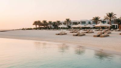 Al Zorah Beach in Ajman at golden hour: soft white sand sweeps into shallow turquoise Gulf water, with a low-rise modern beach resort visible in the deep background framed by palms and a few empty parasols on the upper sand