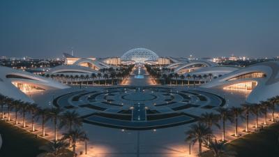 Expo City Dubai pavilions complex at evening with futuristic curving white-and-tan architectural pavilions, palm-lined walkways, and the Al Wasl Plaza dome silhouette in the background