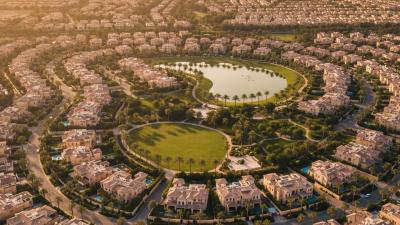 Aerial view of a master-planned Dubai family villa community at golden hour with terracotta-tiled roofs, palm-lined streets, a community park and lake, and the Burj Khalifa silhouetted in the deep distance