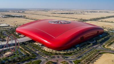 Aerial view of Ferrari World Abu Dhabi on Yas Island: the unmistakable bright red curving roof in the shape of a Ferrari GT car body dominates the frame, with manicured palm-lined paths and access roads spreading out across the desert sand and a roller-coaster track loop visible at the edge