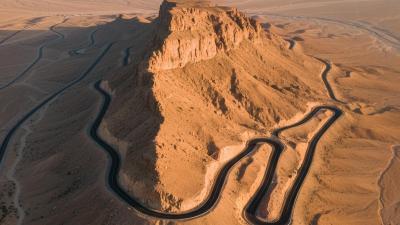 Aerial view of the Jebel Hafeet mountain road in Al Ain at golden hour: a smooth black tarmac road snakes through dramatic switchbacks down the side of an isolated limestone massif rising abruptly from the flat desert below, warm orange and ochre rocky cliffs catching the late-afternoon light