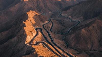 Jebel Jais Mountain Road in Ras Al Khaimah at golden hour: a smooth black tarmac road curves through stark sun-warmed limestone cliffs and rocky escarpments of the Hajar Mountains, with multiple switchbacks visible cascading down the slopes in warm orange and ochre tones