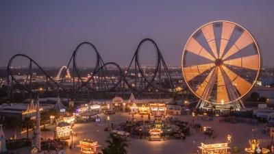 Twilight view of a Yas-Island-style theme park with a looping steel roller coaster silhouette, an illuminated Ferris wheel with motion-trail lights, and a glowing central plaza