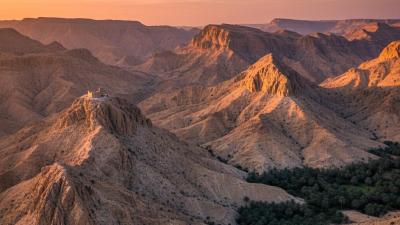 Sweeping landscape of the Hajar Mountains around Masfout in Ajman at sunset: stark sun-warmed beige and ochre limestone cliffs and ridges with deep V-shaped valleys, a small traditional hilltop fort silhouetted on a rocky outcrop and date-palm groves on the valley floor