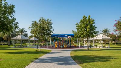 Mushrif Central Park in Abu Dhabi: a wide paved family path winds between large grass picnic lawns and clusters of shade gazebos, with a colourful children's playground mid-frame under mature shade trees