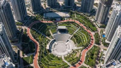 Aerial view of Reem Central Park on Al Reem Island in Abu Dhabi: a paved running loop winds through landscaped lawns, a concrete skate bowl, and a stepped amphitheatre, surrounded by a tight ring of contemporary glass-and-steel residential towers under various stages of completion