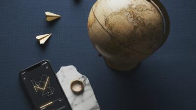 Conceptual editorial overhead photograph on deep navy linen of a wooden world globe tilted slightly off-centre, three small brass paper-aeroplane shapes flying in an arc toward it, and a sleek smartphone showing an abstract transfer-confirmation interface in geometric shapes