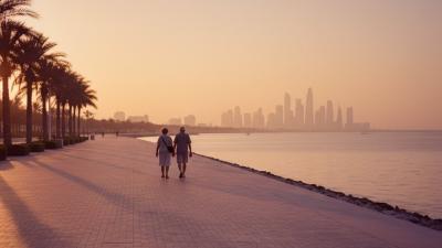 Wide editorial view at sunset along a quiet Saadiyat-style UAE corniche promenade with silhouettes of an older couple walking slowly side-by-side ahead in the frame, faces turned away, the Arabian Gulf glowing peach and gold to their right and low coastal palms casting long shadows
