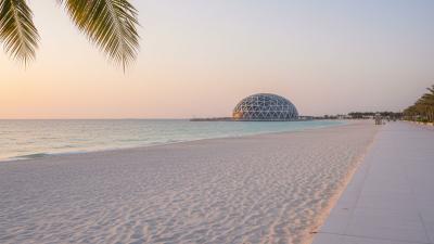 Late-afternoon view across Saadiyat Beach in Abu Dhabi: a paved promenade and palm fronds frame the right side of the image, with the silver geodesic dome of the Louvre Abu Dhabi visible low on the horizon across the white sand and turquoise Gulf water