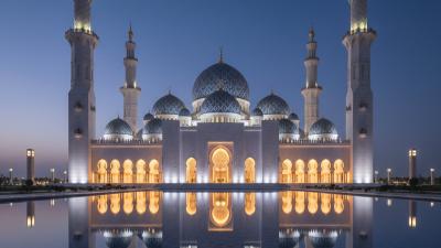 Sheikh Zayed Mosque Ajman at blue hour: a modern white-marble mosque exterior with distinctive blue-and-gold domes and two tall slender minarets, subtly uplit against a deep twilight indigo sky reflected in a foreground pool