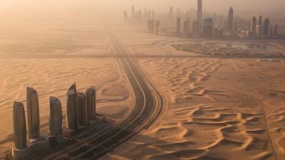Aerial view at golden hour of a UAE highway curving through pale desert dunes — a small cluster of contemporary Abu Dhabi towers on the left and the Dubai skyline with the Burj Khalifa silhouette in the warm haze on the right.