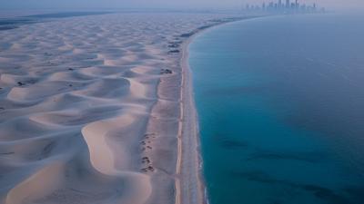 Aerial dawn view of the UAE coastline — pale sand dunes meeting the turquoise Persian Gulf, with the Dubai skyline softly visible in the distant blue-hour haze.