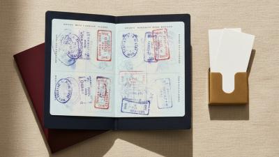 Overhead flat lay of an open passport showing pages stamped with abstract travel stamps in blue and red, a closed burgundy passport stacked beneath, and a brass card holder with two folded paper slips on a beige linen surface.