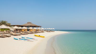 Yas Beach on Yas Island, Abu Dhabi: a long curve of soft white sand meets calm turquoise Arabian Gulf water, with stand-up paddleboards and a kayak resting on the sand and rows of beige cushioned sunbeds with white parasols beneath a palm-thatched bar pavilion