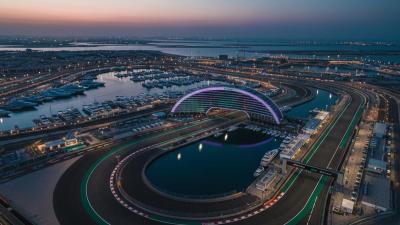 Aerial dusk view of Yas Marina F1 Circuit in Abu Dhabi: the snaking black asphalt race track winds through the frame with the curving illuminated W Abu Dhabi hotel arc straddling the track in the centre, glowing soft purple and teal LED, and the marina with moored superyachts alongside