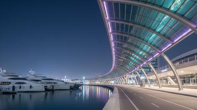 Yas Marina Walk at night: the curved illuminated arc of the Yas Hotel glows soft purple and teal LED light over the running path, with moored superyachts in the marina basin reflecting the colours on the dark water
