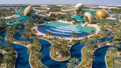 Aerial view of Yas Waterworld Abu Dhabi: vibrant blue and turquoise lazy-river channels and pools wind between curving fibreglass slide structures themed in pearl-diving aesthetics, with a central wave pool and palms throughout