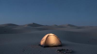 Lit dome tent on rippled desert sand at blue hour with dune ridges receding behind