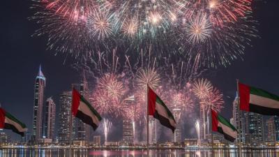 Fireworks in UAE flag colours over a corniche skyline lined with UAE flags on a public holiday night