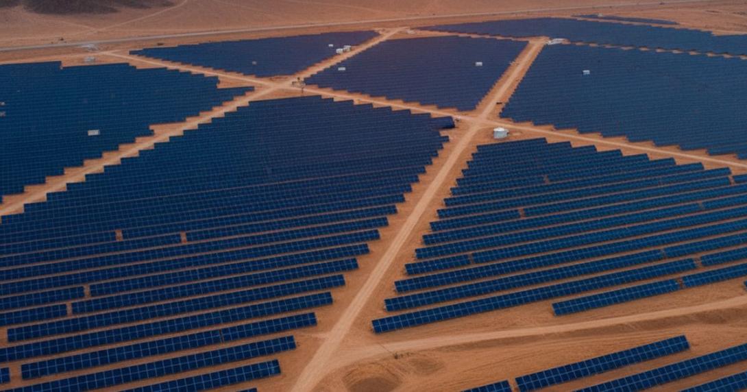 Aerial view of a vast desert solar farm at golden hour with thousands of dark blue solar panels in geometric rows stretching to the horizon and rocky mountains in the deep background
