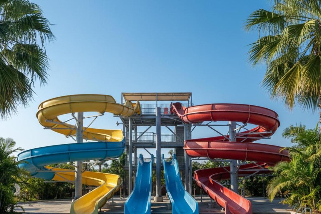 Tall colourful waterpark slide complex at Dreamland Aqua Park in Umm Al Quwain: yellow, blue, and red curving fibreglass slides twist down a tall steel tower into turquoise pools, framed by green palms against a clear blue sky