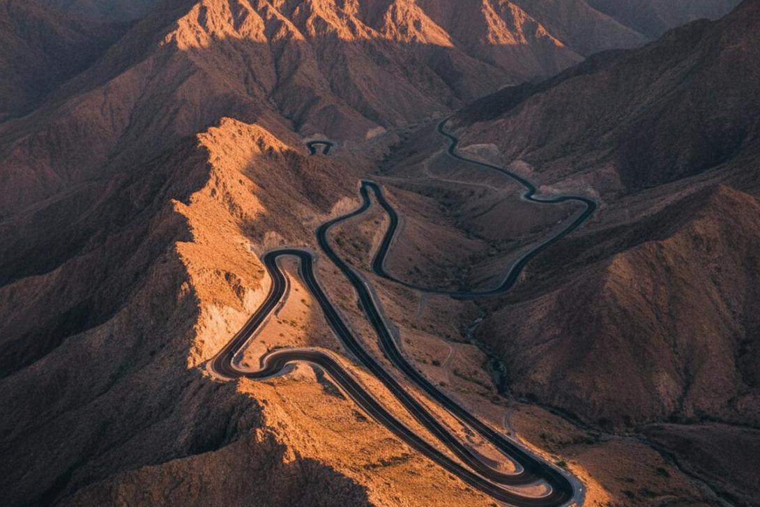 Jebel Jais Mountain Road in Ras Al Khaimah at golden hour: a smooth black tarmac road curves through stark sun-warmed limestone cliffs and rocky escarpments of the Hajar Mountains, with multiple switchbacks visible cascading down the slopes in warm orange and ochre tones