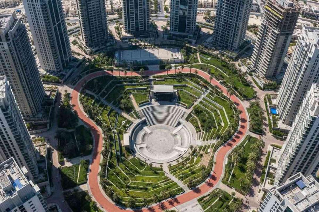 Aerial view of Reem Central Park on Al Reem Island in Abu Dhabi: a paved running loop winds through landscaped lawns, a concrete skate bowl, and a stepped amphitheatre, surrounded by a tight ring of contemporary glass-and-steel residential towers under various stages of completion