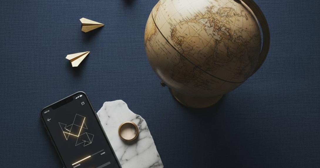 Conceptual editorial overhead photograph on deep navy linen of a wooden world globe tilted slightly off-centre, three small brass paper-aeroplane shapes flying in an arc toward it, and a sleek smartphone showing an abstract transfer-confirmation interface in geometric shapes
