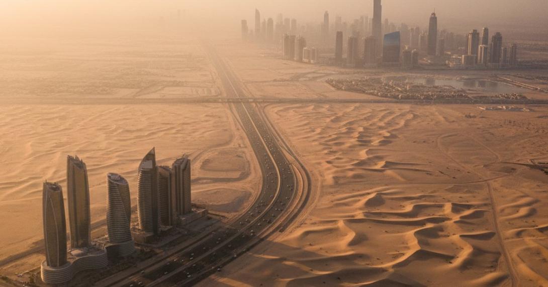 Aerial view at golden hour of a UAE highway curving through pale desert dunes — a small cluster of contemporary Abu Dhabi towers on the left and the Dubai skyline with the Burj Khalifa silhouette in the warm haze on the right.