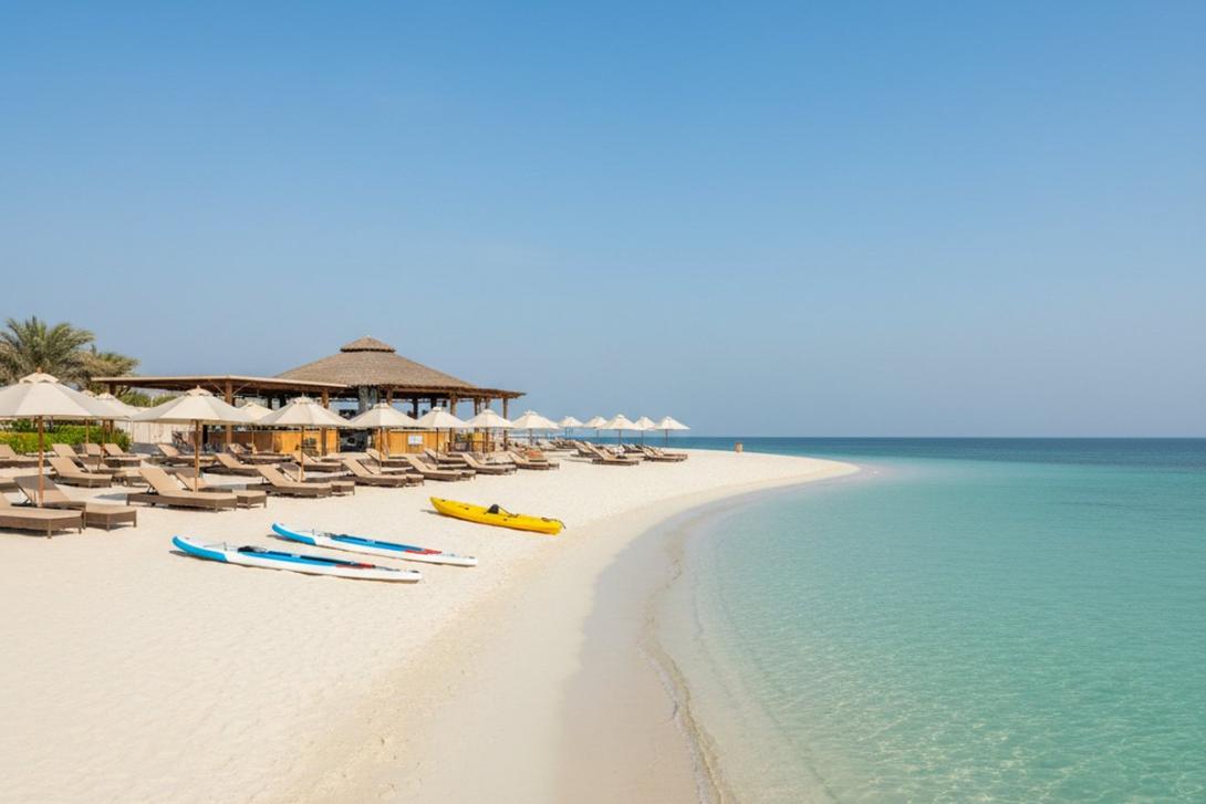 Yas Beach on Yas Island, Abu Dhabi: a long curve of soft white sand meets calm turquoise Arabian Gulf water, with stand-up paddleboards and a kayak resting on the sand and rows of beige cushioned sunbeds with white parasols beneath a palm-thatched bar pavilion