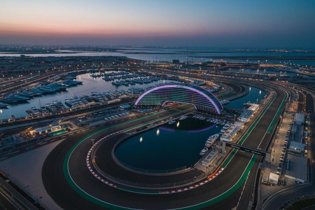 Aerial dusk view of Yas Marina F1 Circuit in Abu Dhabi: the snaking black asphalt race track winds through the frame with the curving illuminated W Abu Dhabi hotel arc straddling the track in the centre, glowing soft purple and teal LED, and the marina with moored superyachts alongside